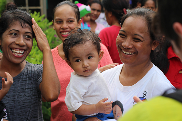 A woman holds her child and smiles at her surrounded by smiling women. 